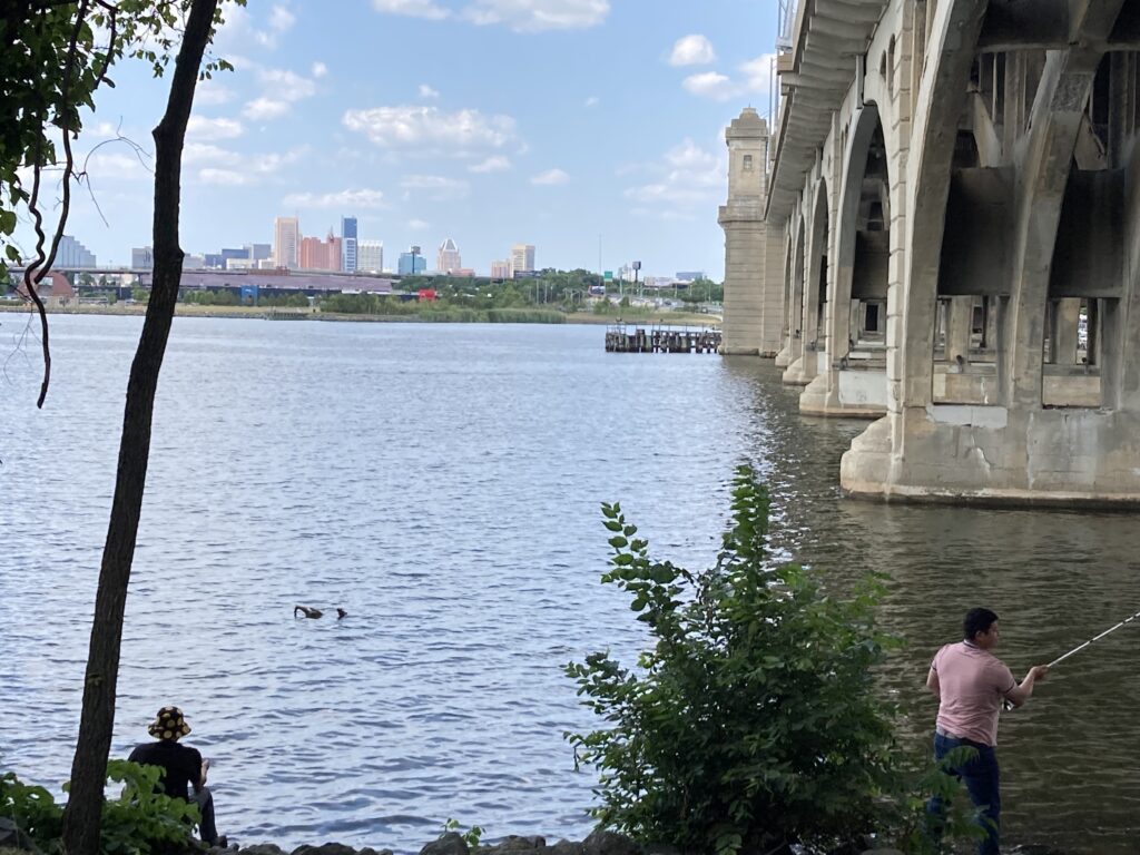 A view of the undeveloped shoreline of the middle branch of the Patapsco River where people hangout for recreational activities, with the historical Hanover Street Bridge overhead and the Baltimore skyline in the background. Photo: Norman Gomlak / The Baltimore Banner.