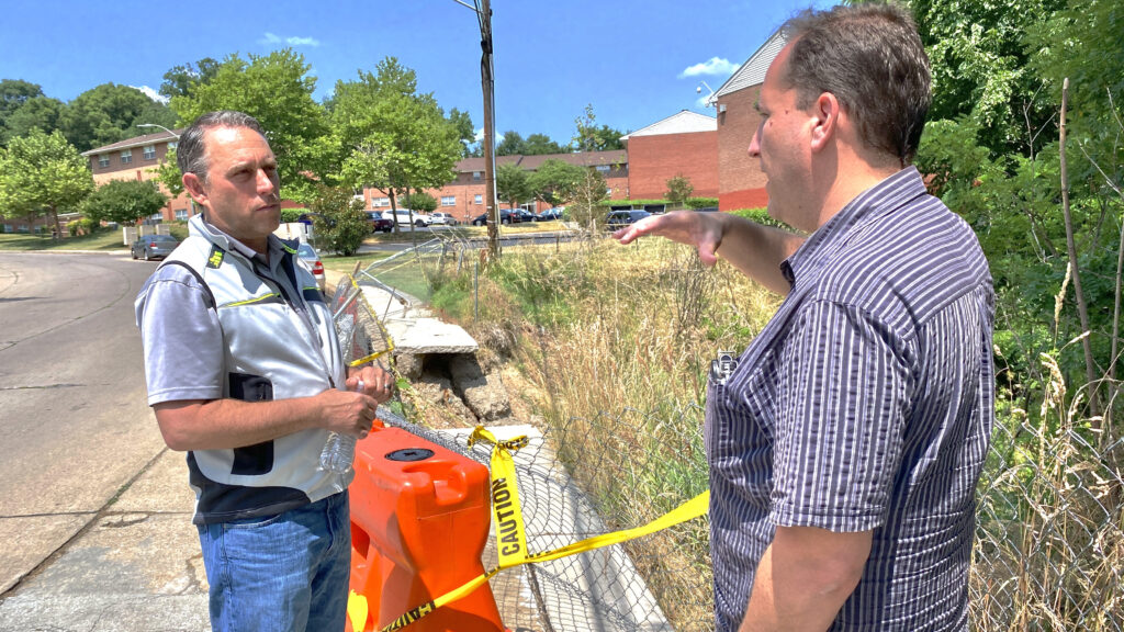 Standing beside a dilapidated road next to a creek, Brad Rogers, right,  environmental consultant Bret Berkley discuss pitching a project to the U.S. Department of Transportation about protecting the road infrastructure in the Cherry Hill neighborhood from storm surges and flooding. Credit: Aman Azhar / Inside Climate News